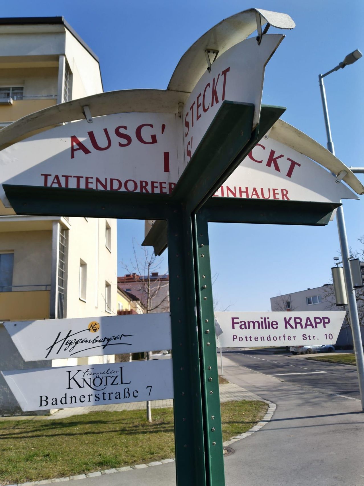 Ein Straßenschild mit drei Pfeilen, die auf verschiedene Straßen zeigen: Ausg, Tattendorfer und Stecket. Darunter befinden sich zwei Schilder für Familien Knottl und Heggamberger. Im Hintergrund sind Gebäude und eine Straße mit geparkten Autos zu sehen.