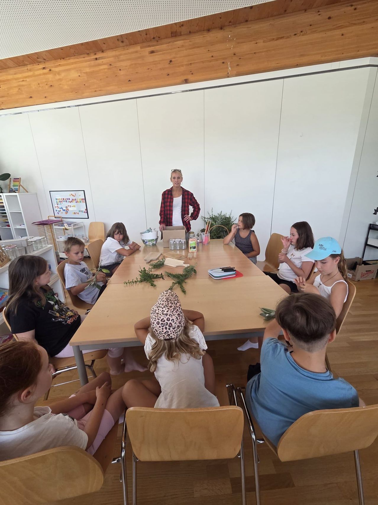 Eine Frau steht am Kopfende eines langen Tisches, umgeben von Kindern, die an einer Aktivität teilnehmen. Der Tisch ist mit Pflanzen, Bechern und anderen Gegenständen gedeckt. Die Kinder sitzen in Stühlen um den Tisch herum und beobachten aufmerksam die Frau.