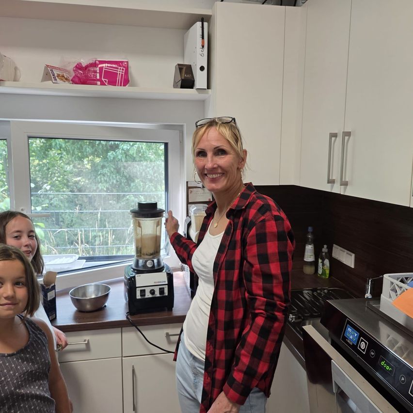 Eine Frau in einem rot-schwarzen Karohemd steht in einer Küche mit zwei jungen Mädchen, eine hält einen Mixer. Hinter ihr ein Fenster mit Bäumen draußen.