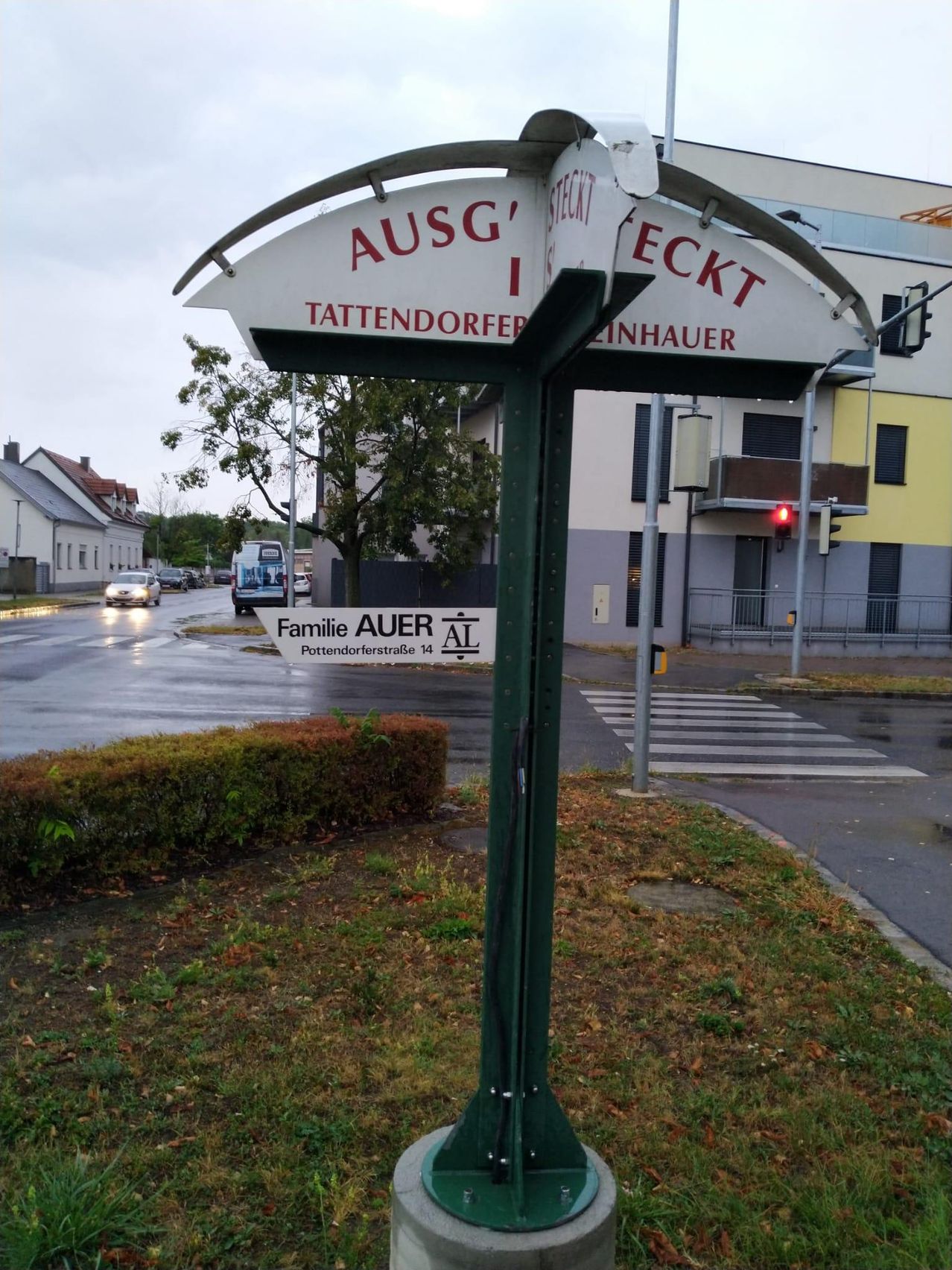 Ein Straßenschild in Deutschland zeigt den Straßennamen und einen Familiennamen an, mit einem Gebäude und einem Zebrastreifen im Hintergrund.