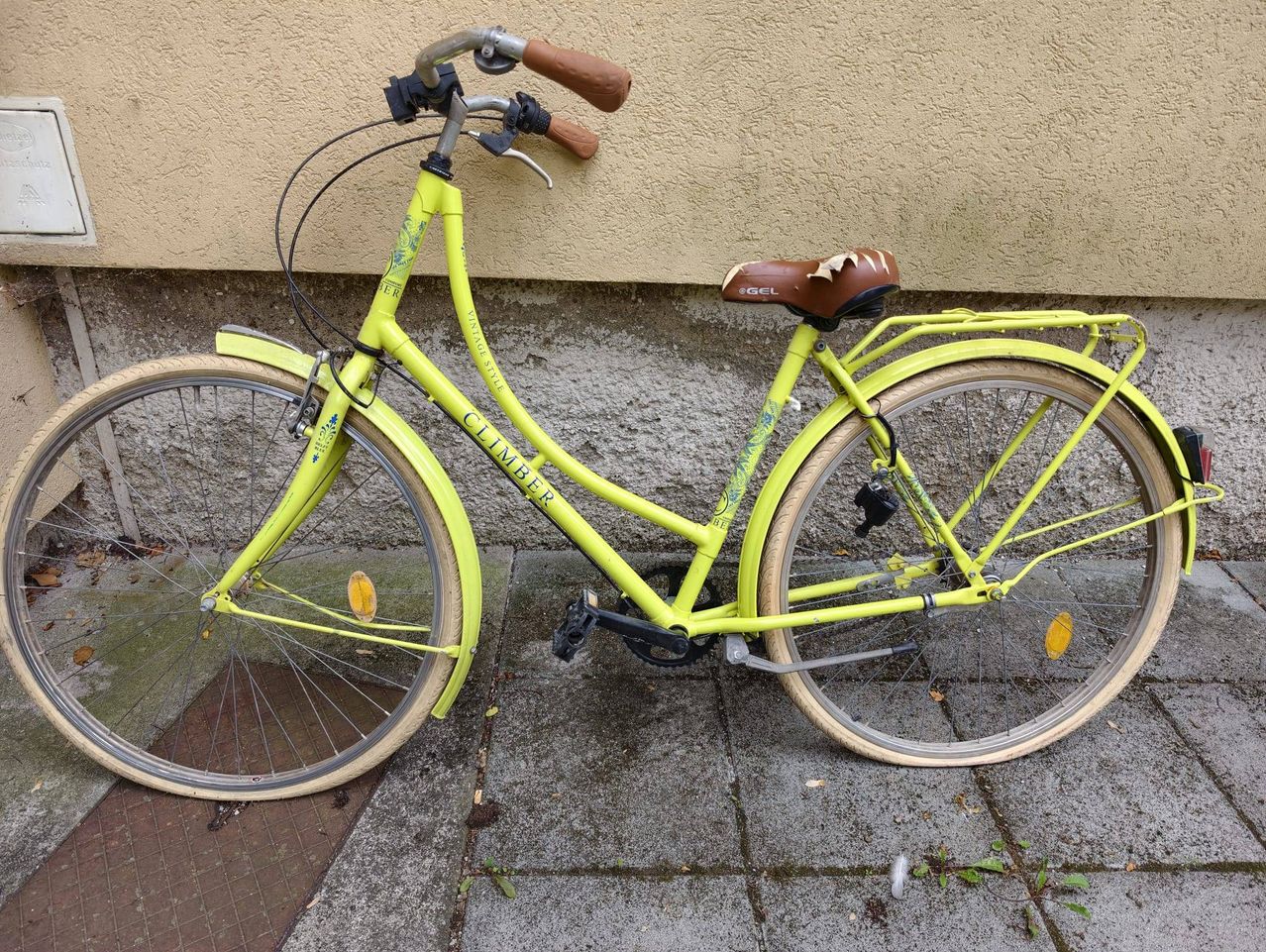 Ein gelbes Vintage-Fahrrad mit braunen Lenkergriffen und einem braunen Sattel steht gegen eine strukturierte Wand gelehnt. Das Fahrrad hat einen Heckträger und Reflektoren.