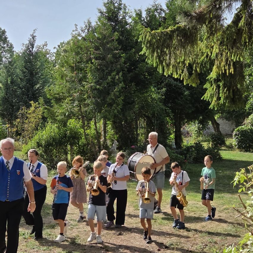 Eine Gruppe von Kindern und Erwachsenen spielt Musikinstrumente in einem Park. Sie stehen in einer Reihe, einige halten Trompeten, andere Posaunen und einige halten Trommeln. Die Erwachsenen tragen Westen und die Kinder tragen Freizeitkleidung. Der Park ist von Bäumen und Gras umgeben.