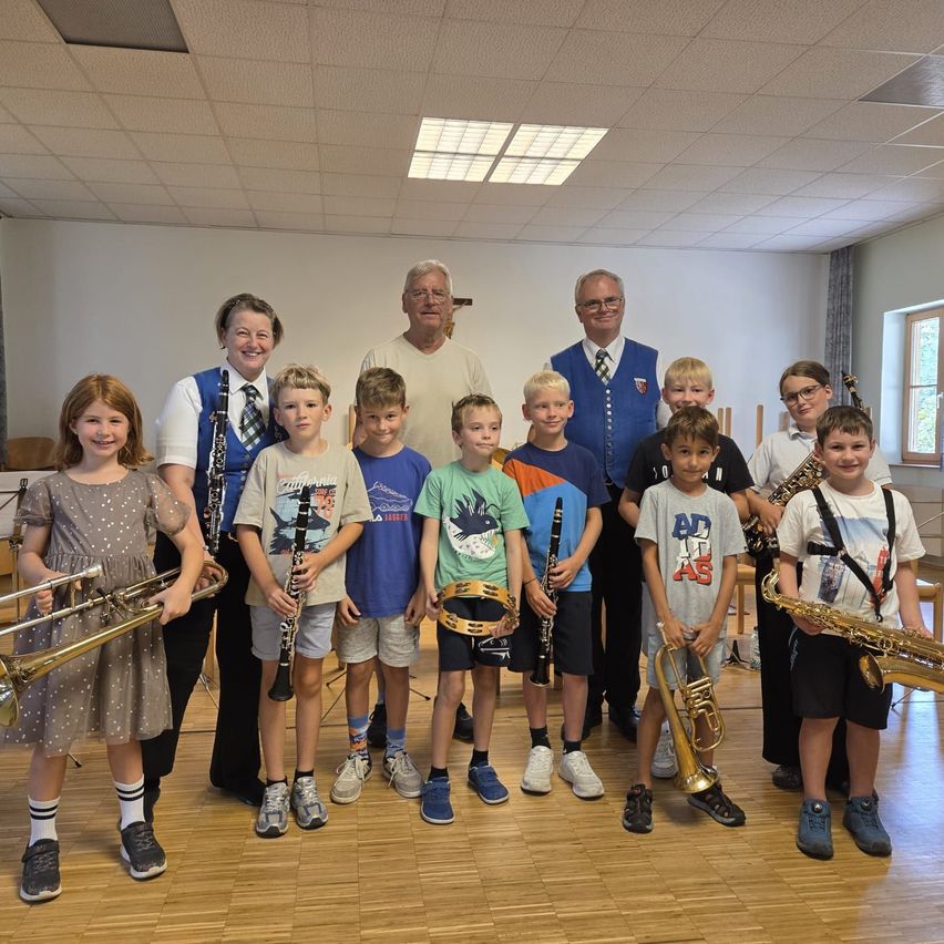 Eine Gruppe von Kindern und Erwachsenen posiert für ein Foto und hält verschiedene Musikinstrumente. Die Kinder halten Trompeten, Posaunen und Klarinetten, während die Erwachsenen Saxophone halten. Sie stehen in einem Raum mit Holzböden und weißen Wänden.