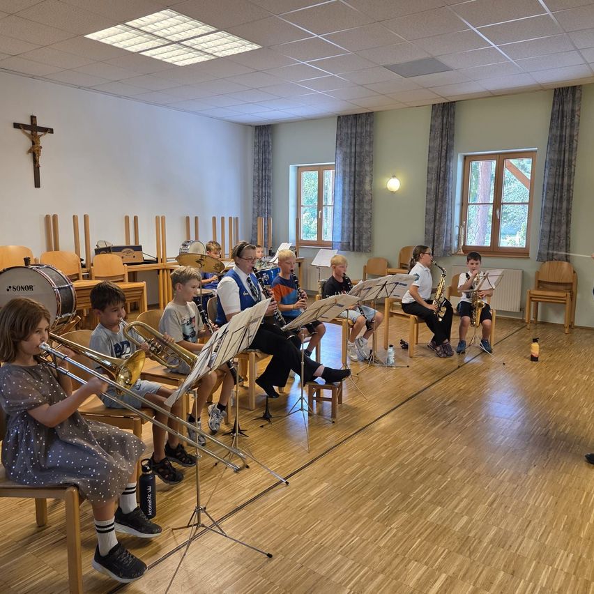 Eine Gruppe von Kindern und Erwachsenen übt Musik in einem Raum mit Holzböden. Sie spielen verschiedene Instrumente wie Trompeten, Posaunen und Saxophone. Eine Person steht vor ihnen, möglicherweise ein Lehrer.