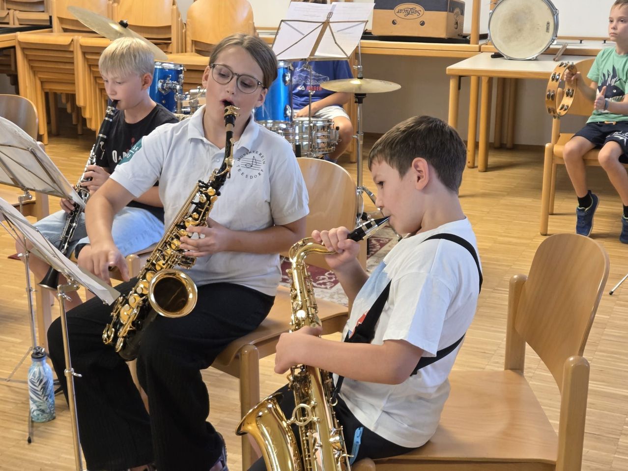 Drei junge Jungen sitzen auf Stühlen und spielen in einem Klassenzimmer Saxophon. Der Junge auf der linken Seite spielt Klarinette, während der Junge in der Mitte Saxophon spielt. Der Junge auf der rechten Seite spielt Saxophon und trägt ein weißes Hemd.