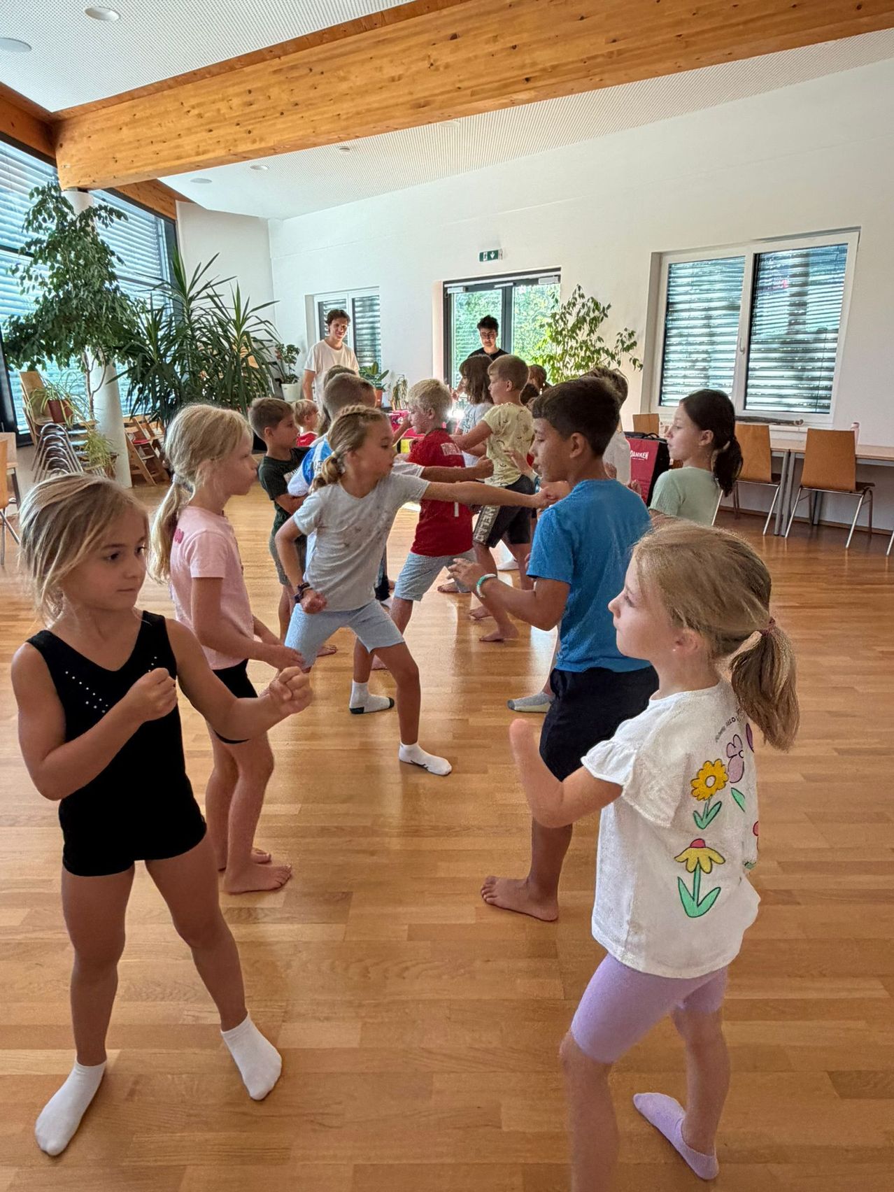 Eine Gruppe von Kindern übt Kampfkunst in einem Raum mit Holzböden und großen Fenstern.