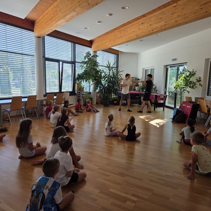 Eine Gruppe von Kindern sitzt auf dem Boden in einem Raum mit Holzböden, während zwei Erwachsene stehend in der Mitte sprechen. Der Raum hat große Fenster mit Jalousien und Topfpflanzen.