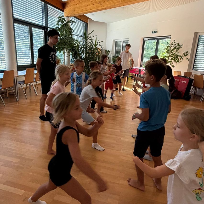 Eine Gruppe von Kindern tanzt in einem Raum mit Holzboden, zwei Erwachsene beaufsichtigen.