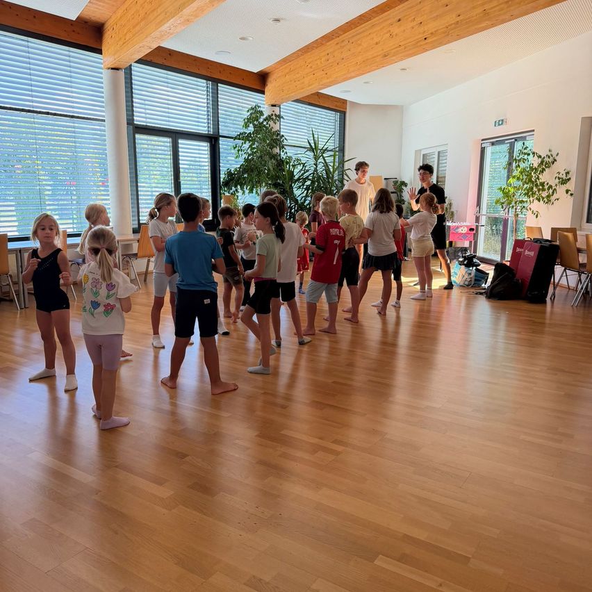 Eine Gruppe von Kindern und zwei Erwachsenen stehen in einem Raum mit Holzböden, großen Fenstern und Pflanzen. Die Kinder sind barfuß und scheinen an einem Tanz- oder Bewegungskurs teilzunehmen.