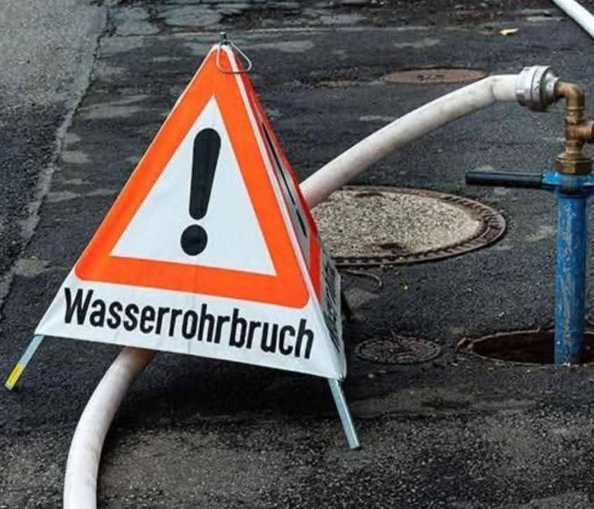 Ein Schild mit Ausrufezeichen und dem Text 'Wasserrohrbruch' steht neben einer gebrochenen Wasserleitung auf dem Boden.