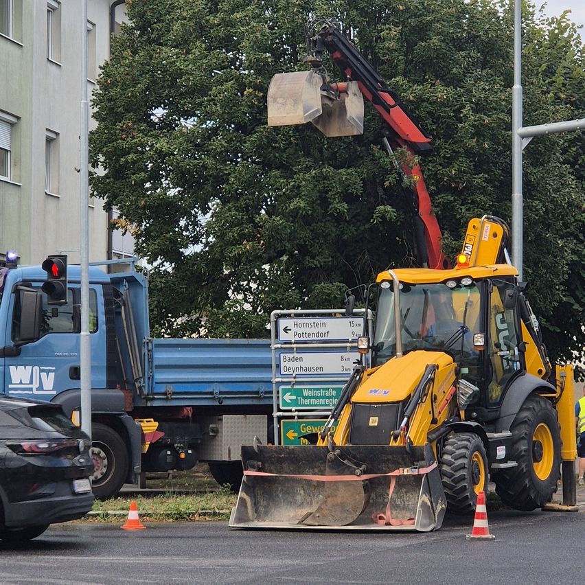 Ein gelber Bagger steht am Straßenrand neben einem blauen Lastwagen und einem Verkehrskegel. Der Bagger ist mit einem Eimer und einem Schaufel ausgestattet.