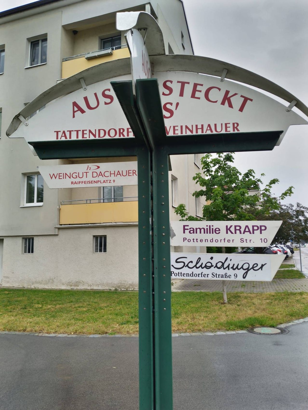 Straßenschild mit mehreren Namen, darunter 'Steckt', 'Tattendorf', 'Weingut Dachauer', 'Familie KRAPP' und 'Schösdiinger'. Dahinter befindet sich ein Gebäude mit Fenstern, einem Balkon und geparkten Autos.