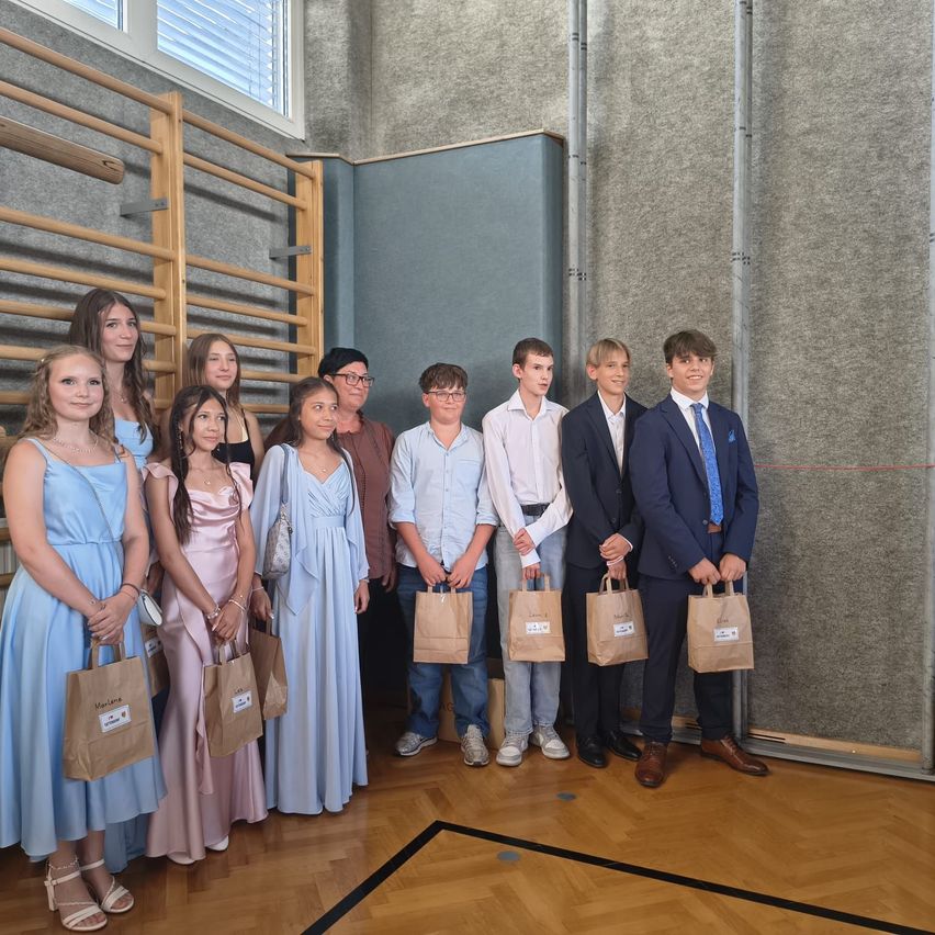 A group of students, both male and female, stand together holding paper bags, likely celebrating an event. The room has wooden floors and a window with blinds.
