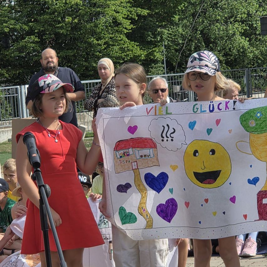 Two young girls hold a handmade sign with drawings and text, standing at a microphone, with onlookers behind them.