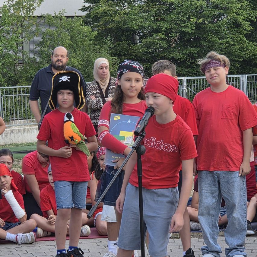 A group of children in red shirts and headbands are standing on a stage. One child is holding a parrot toy while another is singing into a microphone. Behind them, a woman and a man are watching.
