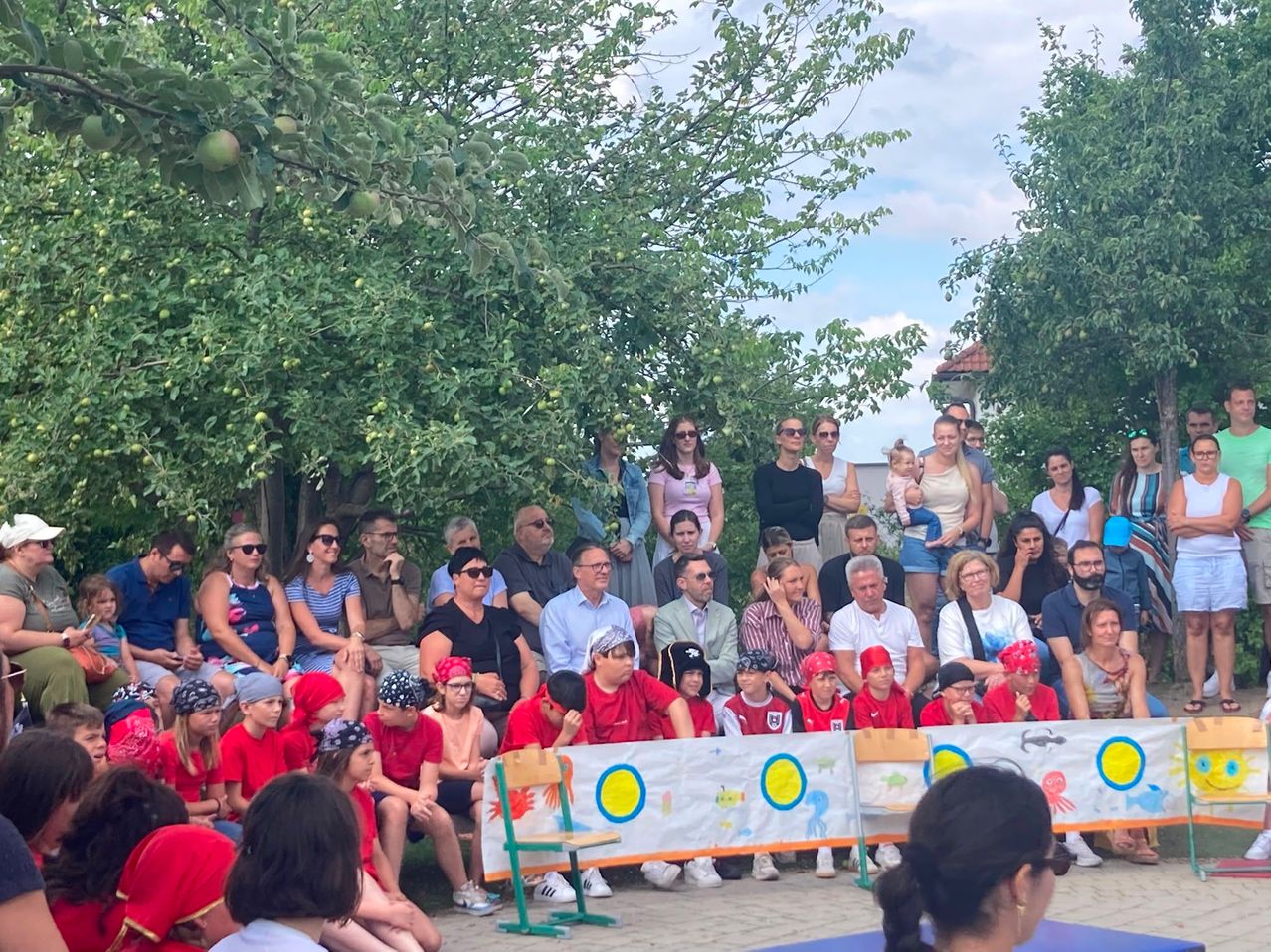 A group of people in red hats and shirts gathered outdoors under trees. Some are sitting while others stand. A banner with designs is in front.
