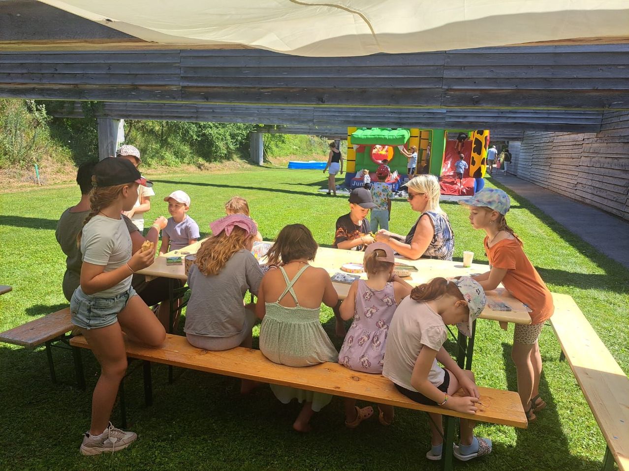 Eine Gruppe von Kindern und Erwachsenen sitzt an einem Picknicktisch in einem Park. Ein Kind isst, während andere den Kopf senken. Eine Frau spricht mit einem Kind. Im Hintergrund sind ein aufblasbarer Rutschvergnügen und ein Hüpfburg zu sehen.