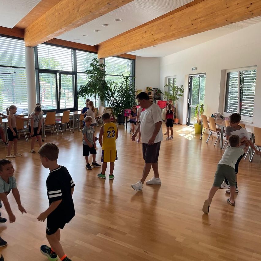 Eine Gruppe von Kindern spielt Basketball in einer Halle mit Holzböden. Ein Mann steht in der Mitte, und andere beobachten in der Nähe. Es gibt große Fenster und Topfpflanzen.