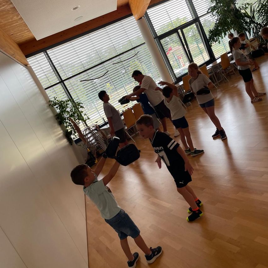 Eine Gruppe von Kindern übt Boxen in einem Raum mit Holzböden. Zwei Kinder halten schwarze Boxhandschuhe. Der Raum hat Glasfenster mit Jalousien und außen stehen Topfpflanzen.
