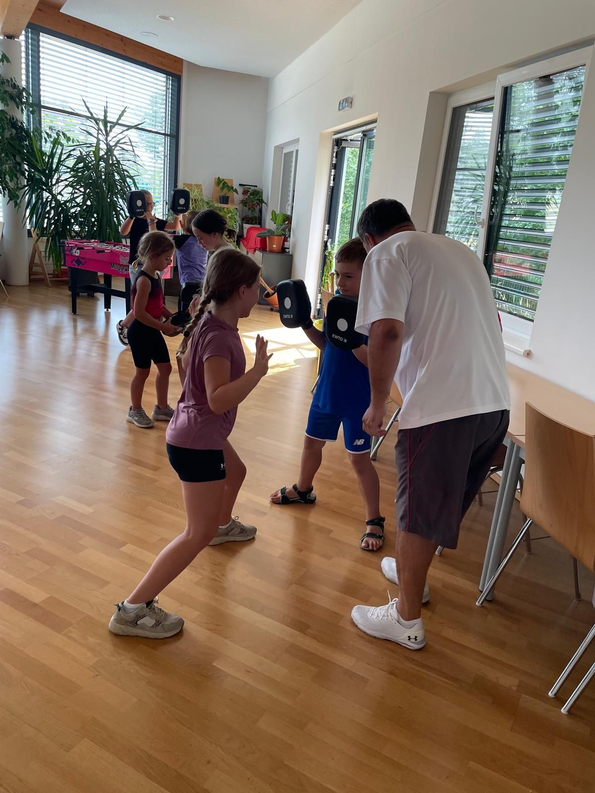 Eine Gruppe von Kindern übt Boxen in einem Raum. Ein Mann coacht sie, und sie tragen Boxhandschuhe. Sie stehen auf einem Holzboden in der Nähe eines Tisches und einer Topfpflanze.