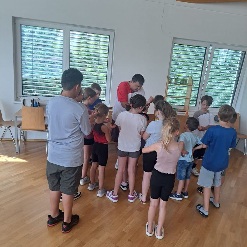 Eine Gruppe von Kindern und zwei Erwachsenen steht in einem Kreis in einem Raum. Ein Erwachsener trägt eine rote Bandana. Der Raum hat Holzböden, weiße Wände und große Fenster mit Jalousien.