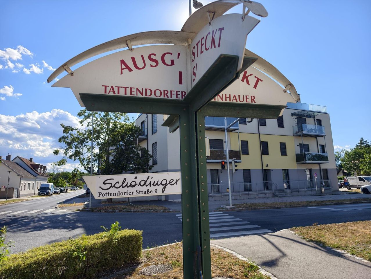 Ein Straßenschild weist auf 'Ausgang Tattendorfer' und 'Steckt S' 'Inhauer' hin, das Schild darunter zeigt 'Schlödiger Pottendorfer Straße 9'. Im Hintergrund sind ein Gebäude und Bäume zu sehen.
