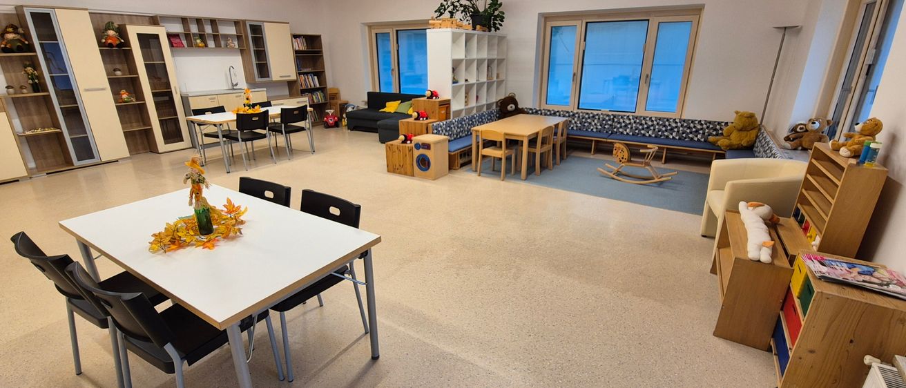 Ein Raum mit Holzdecke und weißen Wänden. Es gibt einen Esstisch, Stühle, eine Couch, Bücher, ein Schaukelpferd und einen Teddybären.