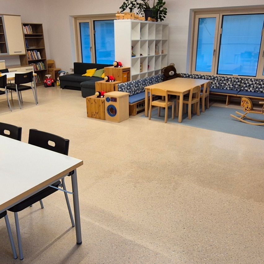 Ein Raum mit Holzdecke und weißen Wänden. Es gibt einen Esstisch, Stühle, eine Couch, Bücher, ein Schaukelpferd und einen Teddybären.