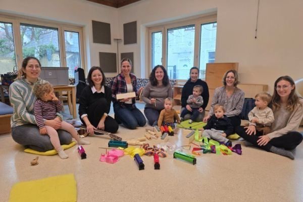 Eine Gruppe von Frauen und Kindern sitzt auf dem Boden in einem Raum mit Musikinstrumenten und lächelt sich gegenseitig an.