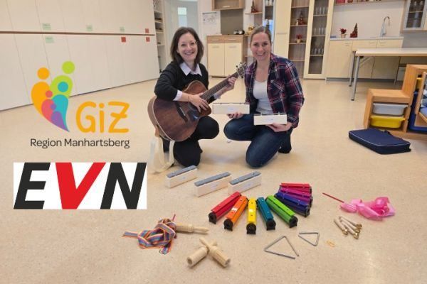 Zwei Frauen lächeln, während sie auf dem Boden knien. Eine hält eine Gitarre und die andere eine Schachtel. Musikinstrumente sind um sie herum verstreut. Das Bild ist mit Giz und EVN beschriftet.