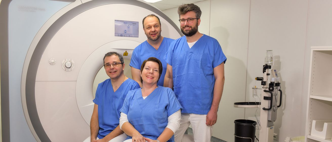 Vier medizinische Mitarbeiter lächeln vor einem MRT-Gerät in einem medizinischen Raum.