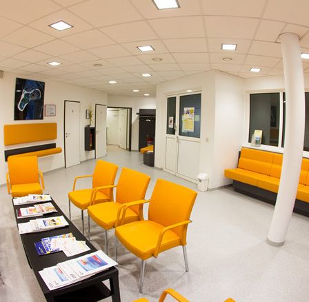 Ein Wartezimmer mit gelben Stühlen, einem Schreibtisch mit Papieren und einer großen Säule.