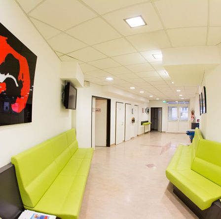 Ein breiter Flur mit weißen Wänden und einem roten abstrakten Gemälde auf der linken Seite. Grüne Sofas mit Zeitschriften stehen auf beiden Seiten des Flurs.