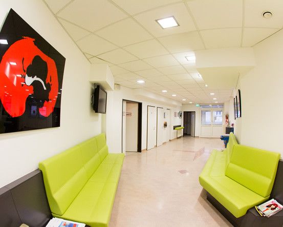 Ein breiter Flur mit weißen Wänden und einem roten abstrakten Gemälde auf der linken Seite. Grüne Sofas mit Zeitschriften stehen auf beiden Seiten des Flurs.