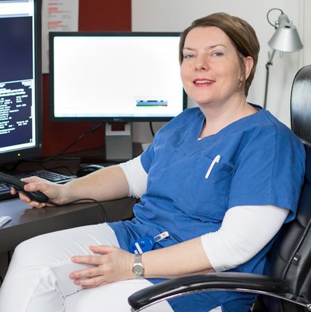 Eine lächelnde Frau in einer blauen Uniform sitzt in einem Bürostuhl mit einer Fernbedienung in der Hand, mit zwei Monitoren und einer Tastatur vor ihr.