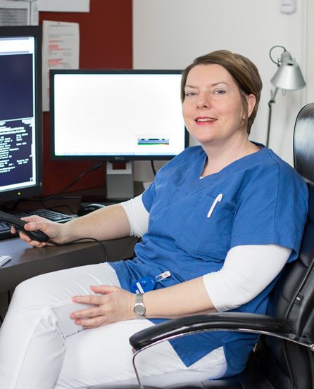 Eine lächelnde Frau in einer blauen Uniform sitzt in einem Bürostuhl mit einer Fernbedienung in der Hand, mit zwei Monitoren und einer Tastatur vor ihr.