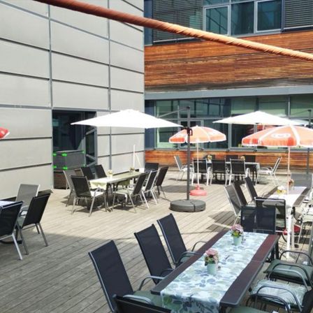 Eine Terrasse mit mehreren Tischen und Stühlen, einige von weißen und orangefarbenen Sonnenschirmen bedeckt. Die Tische haben Tischdecken und Blumenvasen. Im Hintergrund befindet sich ein Gebäude.