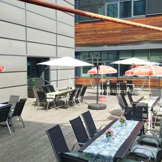 Eine Terrasse mit mehreren Tischen und Stühlen, einige von weißen und orangefarbenen Sonnenschirmen bedeckt. Die Tische haben Tischdecken und Blumenvasen. Im Hintergrund befindet sich ein Gebäude.