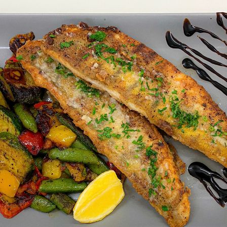 Ein Teller mit gebratenen Fischfilets, garniert mit Petersilie und Zitronenscheiben, begleitet von einer bunten Mischung gerösteter Gemüse.