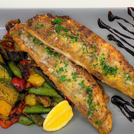 Ein Teller mit gebratenen Fischfilets, garniert mit Petersilie und Zitronenscheiben, begleitet von einer bunten Mischung gerösteter Gemüse.