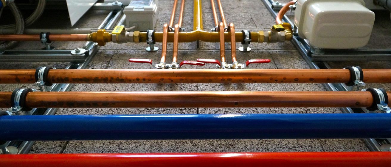 Nahaufnahme von Kupfer- und Stahlrohren mit roten Ventilen und Anschlüssen in einer Anlage. Gelbe, orangefarbene, blaue und rote Rohre sind miteinander verbunden.