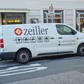 Ein weißer Lieferwagen mit der Aufschrift 'zeiller' und Symbolen für Gas, Wasser, Heizung und Sanitär steht auf der Straße. Dahinter befindet sich ein Gebäude mit einem 30er-Schild.