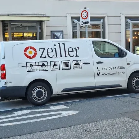 Ein weißer Lieferwagen mit der Aufschrift 'zeiller' und Symbolen für Gas, Wasser, Heizung und Sanitär steht auf der Straße. Dahinter befindet sich ein Gebäude mit einem 30er-Schild.