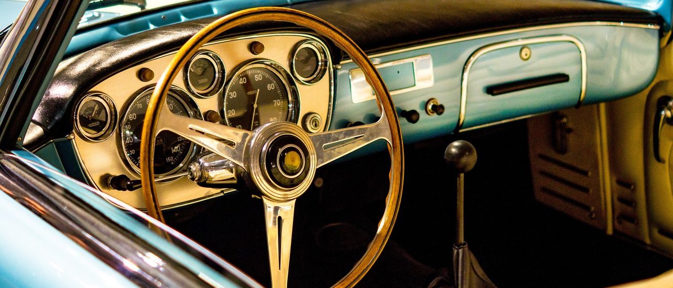 Das Interieur eines Oldtimers zeigt einen Armaturenbrett mit Anzeigen, einem hölzernen Lenkrad und einem Spiegel. Die blauen und beigefarbenen Farben des Autos sind sichtbar.