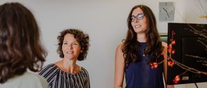 Drei Frauen befinden sich in einem Büro. Eine Frau steht hinter einem Schreibtisch, lächelt und schaut die anderen beiden Frauen an. Eine Uhr und Pflanzen sind im Hintergrund.