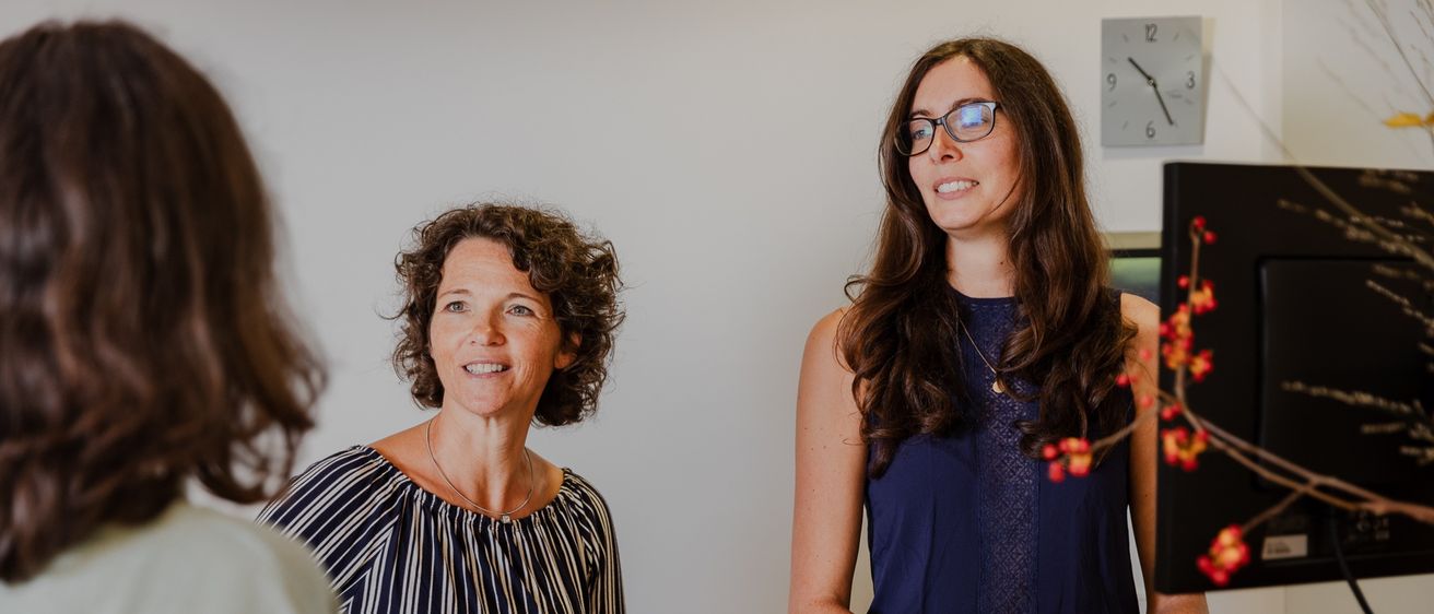 Drei Frauen befinden sich in einem Büro. Eine Frau steht hinter einem Schreibtisch, lächelt und schaut die anderen beiden Frauen an. Eine Uhr und Pflanzen sind im Hintergrund.