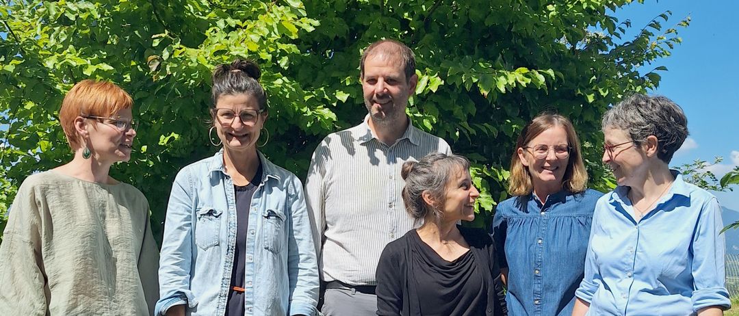 Eine Gruppe von sechs Personen, drei Frauen und drei Männer, stehen gemeinsam vor einem Baum an einem sonnigen Tag.