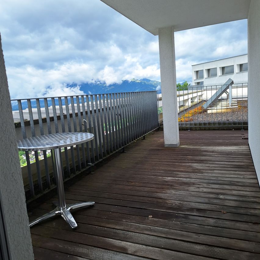 Ein Balkon mit einem Metalltisch und Geländern, mit Blick auf einen bewölkten Himmel und entfernte Berge.