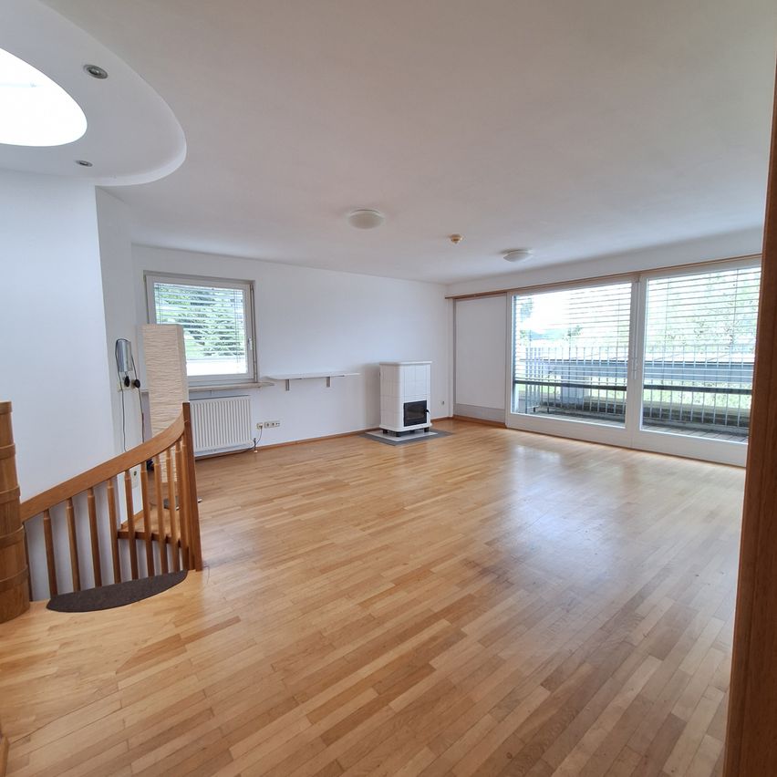 Ein leerer Raum mit Holzböden und weißen Wänden, mit einer Wendeltreppe, einem weißen Heizgerät und großen Fenstern mit Jalousien.