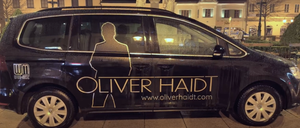 Ein schwarzes Fahrzeug mit goldener Schrift trägt 'OLIVER HAIDT' und 'www.oliverhaidt.com'. Eine Silhouette einer Person ist auf der Seite des Autos. Dahinter steht ein Gebäude mit Statuen.